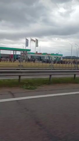 The video shows a view from a vehicle traveling on a highway. On the right side of the highway, there is a gas station with several cars and people present. The sky is overcast with clouds. Further down the road, there are trees and some industrial-looking buildings.