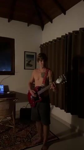 The video shows a young man playing a red electric guitar in a room that appears to be in Goviyapana, Sri Lanka. He is shirtless and wearing dark shorts. He is standing on a patterned rug with a wicker chair and a desk with a laptop to his left. A black amplifier is visible behind him. The room has a wooden ceiling with exposed beams and dark brown curtains covering a window on the right side. A framed picture hangs on the wall. The lighting suggests it is nighttime. The man appears focused on his playing. There is no visible text in the video.