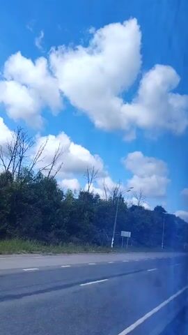 The video shows a highway with a clear blue sky and scattered white clouds. Lush green trees and vegetation line the side of the road. There are streetlights visible along the highway. A white truck and a black car are seen driving on the highway. The camera appears to be moving, possibly from inside a vehicle, as evidenced by the slight blur and the perspective.