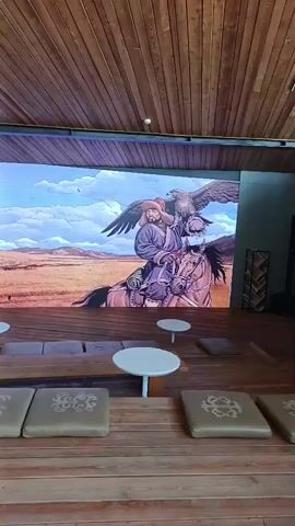 The video shows a wide shot of an indoor space with a wooden ceiling and floor. In the center, there is a display area with a large digital screen showing a rotating series of images. The first image depicts a historical scene with a man on horseback, an eagle, and a pyramid in the background. The subsequent images show a stone structure and a map. In front of the screen, there are rows of cushioned benches and small white tables arranged on a wooden platform. To the left, there is a white podium with a logo that reads "ТУРА-ХАН". The overall environment suggests a museum, exhibition hall, or cultural center.