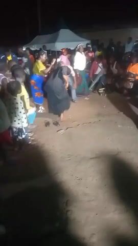 The video shows an outdoor gathering at night. A group of people, including adults and children, are gathered around a central area where two women are dancing. One woman is wearing a black dress, and the other is wearing a white shirt and a green skirt. Several people are seated, watching the dancers. In the background, there are tents and additional spectators. The ground appears to be dirt. The lighting is artificial, suggesting evening or nighttime.