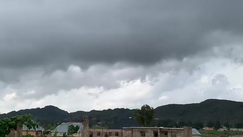 This is a video file of an outdoor scene in Zawan, Nigeria. The sky is overcast with heavy, dark gray clouds, indicating a stormy or rainy day. The time of day appears to be late afternoon or early evening, given the dim light.

In the foreground, there is a partially constructed building with exposed brickwork and concrete pillars. To the left, there are some green plants and a portion of a building with a white roof. Further back, more buildings are visible, some with dark, triangular roofs. A single tall tree with green foliage stands out against the backdrop of rolling hills. The hills themselves are covered in vegetation and appear to be rocky and mountainous.

The overall mood of the scene is somber and dramatic, largely due to the oppressive cloud cover. The atmosphere suggests an impending storm or the aftermath of one. There is no visible text in the media file. The subjects are primarily the natural landscape and the built environment, with no people clearly visible. The activity is passive, capturing a moment in time under a specific weather condition.