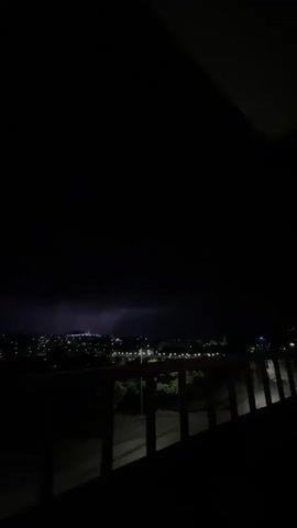 The video shows a night view of a city from a balcony. The city lights are visible in the distance, and a road runs beneath the balcony. A silver car is parked on the road. The balcony has a wooden railing. The sky is dark.