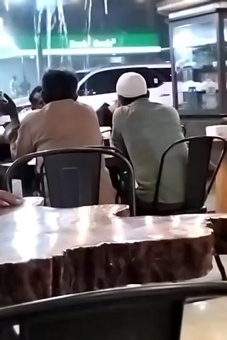 Three men are seated at tables in an indoor or semi-outdoor setting, facing away from the camera. Two of the men are at one table; one man wears a tan shirt and the other a green shirt and a white skullcap. The third man is at a different table partially obscured by a large, shiny, oddly-shaped table in the foreground. The tables are set on what appears to be a patio or porch with a decorative water feature in the background, blurring due to the out-of-focus depth of field. Through the window, the lights and shapes of passing cars are seen on a street.