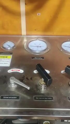 A close-up shot of a stainless steel control panel shows three pressure gauges. The leftmost gauge is partially obscured by a yellow and black warning label that reads "NOTICE" and "DRY". Below the gauges are several levers and knobs. The leftmost control is a silver lever labeled "Driven air switch" with an indicator pointing to "OFF". To its right is a black knob labeled "Gas source input valve" which is in the "OPEN" position, allowing the pressure on the middle gauge to rise from approximately 10 to 20 psi. Further to the right, a portion of another black knob is visible. The background is a textured, wood-like surface.
