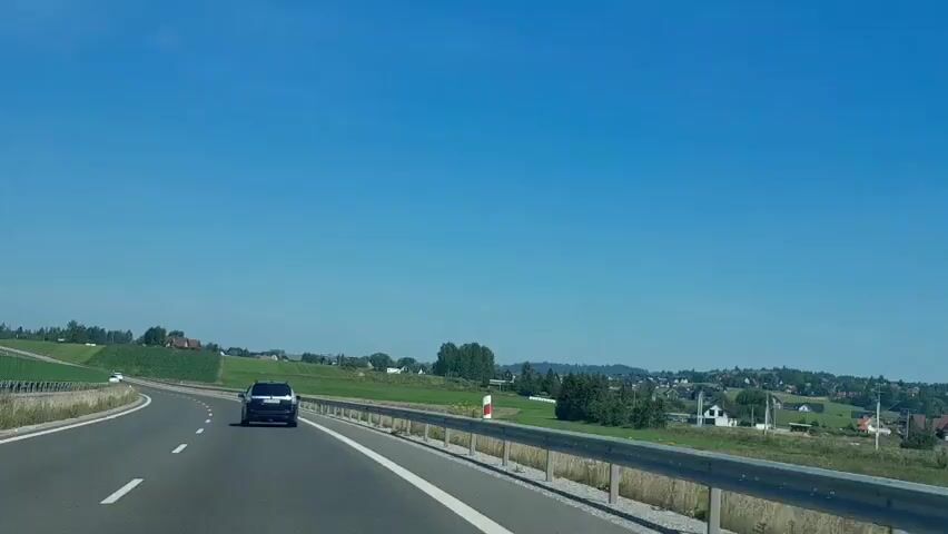 A dark-colored SUV is driving on a highway. The car is in the right lane of a two-lane highway. The highway is curved and goes uphill. The sky is clear and blue. There are green fields and trees on either side of the highway. There are also some houses visible in the distance. The SUV is driving away from the camera.