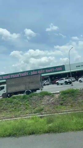 This is a video taken from a moving vehicle, likely a car or bus, on a sunny day in Dong Nai City, Vietnam. The scene is a highway or main road with a rest stop visible in the background. The sky is bright blue with scattered white clouds, indicating fair weather.

Several large trucks are the main subjects in the foreground. One truck, on the left, is a white tanker trailer labeled "SAIGON TANKER TRAILER." To its right, a large truck with a long, flatbed trailer filled with what appear to be stacked wooden planks or logs is moving. Further to the right, another white truck is seen, and a red truck with a trailer is visible in the second clip. The last clip shows a white truck with a large cylindrical tank trailer, labeled "LNG" and "CIMC ENRIC."

In the background, a long building with the sign "FUJITA Rest Stop" is prominent. Below "Rest Stop," there is Vietnamese text that translates to "Diem Den Than Thien" (Friendly Destination). Another sign, partially visible, reads "NGHI PHAN THIET - DAU GIAY Km47+500." This suggests the rest stop is located between Phan Thiet and Dau Giay at kilometer marker 47.5. Several white pickup trucks are parked or driving near the rest stop.

A person in red clothing is visible near the truck on the right in the third and fourth clips, possibly a worker. The overall impression is a typical scene along a busy highway in Vietnam, with transport trucks and service areas. The time of day appears to be daytime, likely late morning or afternoon, given the bright sunlight. There are no overt emotional cues, but the scene conveys a sense of movement and commerce.