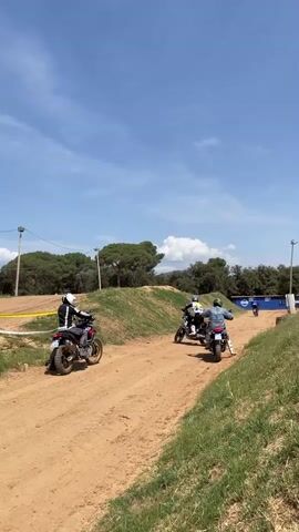 Three people are riding motorcycles on a dirt track on a sunny day. The track is lined with grassy embankments and a yellow caution tape. In the background, there are trees, light poles, and blue containers. The sky is blue with scattered clouds. The motorcycles kick up dust as they drive. The motorcycles drive away from the camera.