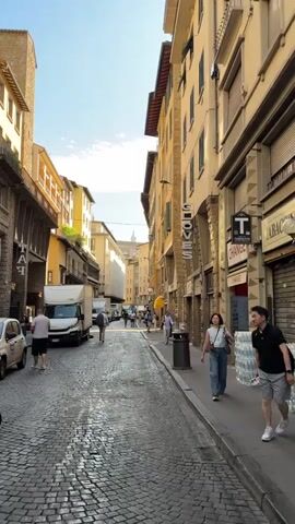 This video captures a street scene in Florence, Italy, likely during the daytime under sunny skies. The street is paved with cobblestones, and tall, historic buildings with multi-story facades line both sides. Many of these buildings appear to house shops, indicated by storefronts and signage.

The primary subjects are people walking and a man riding a bicycle. On the right side of the street, several people are seen walking on the sidewalk. One woman in the foreground wears jeans and a light-colored top, carrying a bag. Further down, other pedestrians are visible. On the left side, a white delivery truck is parked. A man in a red shirt is riding a red and white electric bicycle towards the camera. Another person is walking in the distance on the left.

Notable details include the architecture of the buildings, with their traditional windows and decorative elements. The sunlight casts shadows on the cobblestone street, suggesting it is midday or early afternoon. The general atmosphere seems relaxed and typical of a European city street with pedestrian and vehicle traffic.

Visible text includes "GLOVES" vertically on a sign above a storefront on the right. Another sign on the right reads "TABACCHI" and "CHANGE." On the left side, partially visible text on a building includes "MA" and some numbers. The overall impression is a vibrant, sunlit street in a historic Italian city.