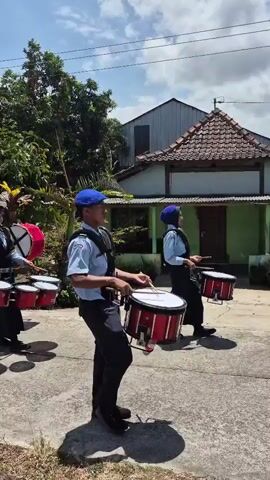 The video shows a marching band performing outdoors. The band members are wearing matching uniforms consisting of blue caps, light blue shirts, and dark pants. They are carrying and playing various drums, including bass drums and snare drums. The band is marching on a paved path, with greenery and houses visible in the background. The sky is blue with some clouds. The camera is positioned to follow the band as they move forward.