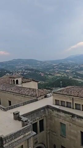 This video shows a panoramic view of a town nestled in rolling hills under a cloudy sky. The foreground features the tiled roofs and stone walls of several buildings, some with visible windows and architectural details. A large, dark green tree dominates the right side of the frame, its branches reaching towards the sky. The landscape beyond the buildings is a patchwork of green fields, scattered houses, and winding roads, with distant mountains fading into the hazy horizon. The overall impression is one of a scenic, historic settlement in a rural, mountainous setting.