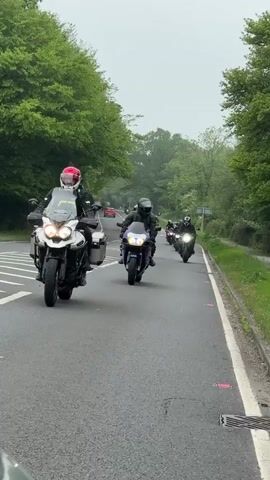 A group of about eight motorcycles is traveling down a gray asphalt road. A white motorcycle with side saddlebags leads the pack. The rider wears a black and white jacket and a red helmet. The second motorcycle is navy blue. Trees border both sides of the road. The weather is cloudy.