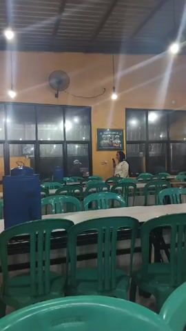 The video shows the interior of a restaurant, possibly a cafeteria or food stall. In the foreground, rows of empty green plastic chairs are visible, neatly arranged around tables covered with white cloths. Behind the tables, a long counter displays various food items in white bowls. A woman wearing a light-colored hijab and a patterned shirt stands behind the counter, facing the camera. To her left, another person is partially visible. Further down the counter, a large silver pot with a lid is present. The walls of the restaurant are painted an ochre color, and the ceiling appears to be made of corrugated metal. Fluorescent lights are installed on the ceiling, casting a bright illumination on the scene. A black oscillating fan is mounted on the wall. The background features large windows, some of which are covered by blinds. There's a sign with text and pictures above the food display. A blue water cooler is positioned to the left of the tables. The overall atmosphere suggests a casual dining establishment.