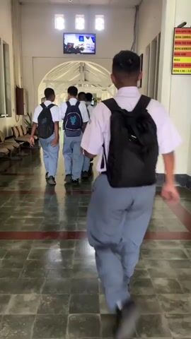 A group of male students wearing white shirts, gray pants, and backpacks are walking down a long, tiled hallway. The hallway has benches on either side and a large arched opening at the far end. A television screen is mounted on the wall above the arched opening. The students are walking away from the camera, with the student in the foreground being the closest to the viewer. The hallway appears to be indoors and well-lit.