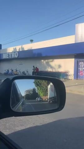 The video shows a view from the driver's side mirror of a car, reflecting a street scene. A large building with a blue and white facade and the word "METRO" in blue letters is visible. Several electric scooters and what appear to be shopping carts are parked outside the building. In the reflection of the mirror, a white truck is driving on the road, along with other cars and buildings. There are trees and power lines visible in the background. The sky is clear and blue. The overall impression is a sunny day in a commercial area.