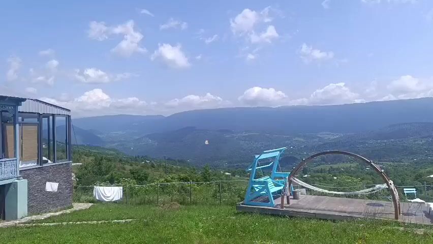 The video shows a panoramic view of a mountainous landscape under a cloudy blue sky. In the foreground, a wooden deck features a turquoise swing set, a hammock suspended from a curved wooden frame, and several white chairs and tables. To the right, a silver minivan is parked on a grassy slope. The background reveals a verdant valley dotted with houses and surrounded by rolling hills and distant mountains. Laundry hangs on a clothesline near a building on the left side of the frame. The camera pans slowly to the right, showcasing the expansive scenery and the recreational area.
