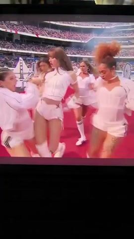 The video displays a performance on a red stage in a large stadium. A central female performer, dressed in white, sings into a microphone, surrounded by five backup dancers also in white outfits. The dancers are performing synchronized choreography. The background shows a blurred stadium audience and a large screen displaying event information. The camera is positioned at a distance, capturing the full stage and performers.