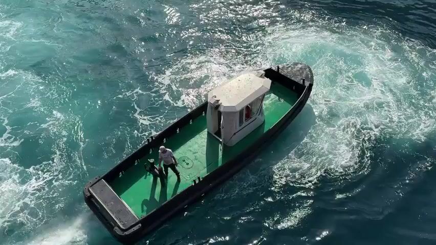 An aerial view shows a small tugboat with a white cabin on a choppy, turquoise body of water. Two people are visible on the deck of the tugboat, which is creating a wake. One person, wearing a light-colored long-sleeved shirt and dark pants, appears to be standing. The other person, wearing dark clothing, is in a squatting or kneeling position. The tugboat is angled, suggesting it is in motion. The water is a vibrant teal with white foam and ripples indicating movement and disturbance.
