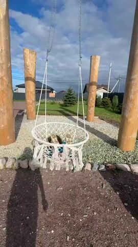 A person is sitting in a hanging swing made of white macrame. The swing is suspended from a wooden A-frame structure composed of four large wooden logs. The swing and the frame are set up in a yard with a gravel ground covering. Behind the swing, there is a lawn with a few small evergreen trees and houses in the background. The sky is blue with scattered clouds. The camera angles shift slightly throughout the video, offering different perspectives of the scene and the person in the swing.