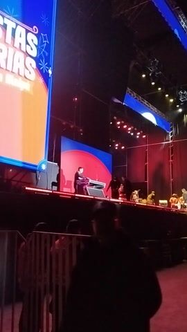 The video captures a dynamic night-time concert or event unfolding on a large, brightly lit stage. In the foreground, one individual is actively engaged with what appears to be a DJ console or sound mixing equipment. Further back on the stage, several other performers are visible; one person is playing a guitar, and another is positioned behind a drum set, indicating a live band performance.

The stage is equipped with elaborate lighting rigs, casting a vibrant array of colors, predominantly red and blue, across the performance area. Large LED screens serve as backdrops, displaying various graphics and text. On one screen, the partial word "FIESTAS" is visible, while another prominently displays "MAIPÚ." The dark surroundings suggest an evening or night event, with the stage lights providing the main illumination. The overall scene conveys the energetic atmosphere of a live musical performance or public celebration taking place in Maipú, Chile.