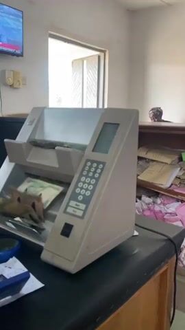 The video shows a cash counting machine in use. The machine is beige and has a keypad and a digital display. Several banknotes are visible inside the machine, being processed. To the left of the machine, a television screen displays news footage of a man in a suit. A window is visible in the background, letting in natural light, suggesting it is daytime. To the right, a shelf is stacked with papers and some pink fabric. A person's head and shoulders are visible in the far right background, looking over the shelf. The scene appears to be an indoor office or administrative setting. The overall impression is one of a functional workspace where cash transactions are being handled. The visible text on the television screen is too blurry to read clearly. The cash counter display shows numbers, indicating the counting process is underway.