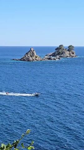 A fast boat with a white hull speeds across the deep blue sea, leaving a foamy wake. The boat appears to be towing a yellow inflatable tube behind it. In the distance, two small, rocky islands rise from the water. One island has a structure resembling a small tower or ruin on its peak, while the other is sparsely covered with green trees. The sky is a clear, bright blue, and the sun appears to be shining, casting a bright light on the scene. The camera is positioned at a high vantage point, looking down on the water.