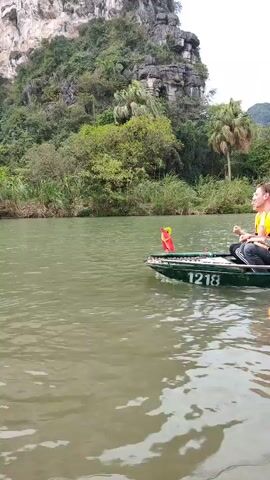 The video shows a small boat traveling on a river against a background of lush green vegetation and a rocky cliff. A person in a yellow life vest is paddling the boat with a long oar. Two other people sit in the back of the boat. The boat is dark green and has the number "1218" painted on the side. A small red Vietnamese flag is attached to the front of the boat. The river is a murky green color, and the vegetation on the riverbank is dense. The cliff is covered in trees and other greenery, and the sky is overcast.