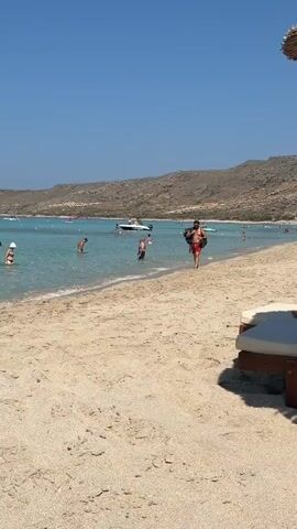 A man walks along a sandy beach towards the camera carrying a large black bag over his shoulder. He is wearing red swimming shorts and is shirtless. In the shallow, clear turquoise water behind him, several people are standing and wading. Further out, a white boat with people on it is anchored. In the background, a rocky, barren hillside rises behind the body of water. The sky is clear and blue.
