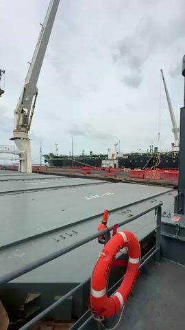 A time-lapse video shows a view from a cargo ship in a port, facing forward and slightly to the right. The ship's deck is visible in the foreground, with two cranes on the left side of the frame. In the distance, other ships are docked or moving in the water. The sky is cloudy. The ship slowly moves forward, revealing more of the port and the other ships. The video ends with the ship still moving forward.
