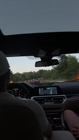 The video shows the interior of a car with a person wearing a white hat driving. The dashboard features a navigation system displaying a map and route. Orange traffic cones are visible on the road ahead, indicating construction or a closed lane. Other cars are driving on the road, and trees line the sides. The sky appears to be overcast. The time stamp is 00:00 to 00:03.