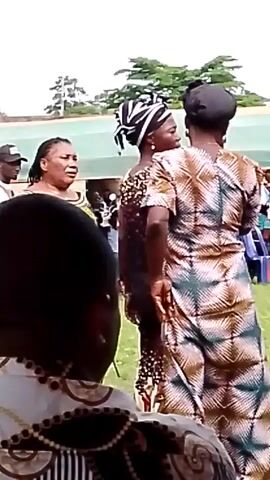 The video shows an outdoor gathering with several people. In the foreground, a person wearing a patterned headdress and top is visible from the back. To their left, a woman with braided hair and a green patterned dress is facing them, speaking. Between these two, another woman with a black headwrap and a black patterned dress is facing away from the camera, looking towards the woman in the green dress. To the right, a woman in a long, multicolored patterned dress with her hands on her hips is facing away from the camera, also engaged in conversation. In the background, more people are seated in chairs under a canopy, and trees and a building are visible. The overall scene suggests a social event or celebration taking place outdoors.