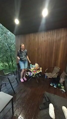 A woman is dancing on a wet wooden porch. She is wearing a t-shirt, denim shorts, and pink flip-flops. She is smiling and has her arms outstretched. A young boy in a red hockey jersey approaches her and holds out a laptop. The woman stops dancing and looks at the boy. There is a table with food and drinks on it behind the woman. Two chairs are visible on the porch. Trees and foliage are visible in the background. The porch is lit by two ceiling lights.
