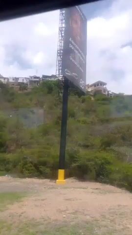 The video shows a view from a moving vehicle, likely a car or bus, looking out a window. The scene is dominated by a lush green hillside covered in dense vegetation. Several houses are visible perched on the top of the hill, appearing somewhat scattered. The foreground shows a dirt and grassy area. The sky is partly cloudy with patches of blue. The footage appears to be taken during the daytime. There are reflections on the window, partially obscuring the view at times. As the vehicle moves, the perspective shifts, revealing more of the hillside and houses. There are some indications of litter or debris on the hillside. The video also briefly shows a dirt road with a few motorcycles parked on it further up the hill, and a concrete structure that might be stairs or a wall.