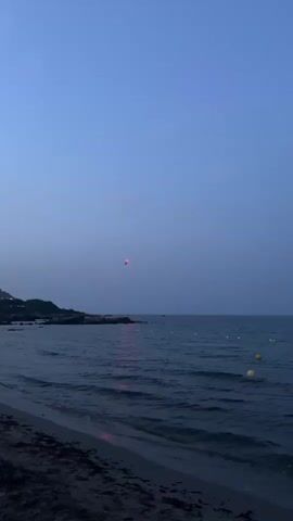 This media file shows a tranquil beach scene at dusk. The sky is a deep, clear blue, fading to a lighter hue near the horizon, suggesting the sun has recently set. Gentle waves lap onto a sandy shore in the foreground, with some seaweed scattered along the waterline.

In the distance, a rocky coastline juts out into the sea, dotted with lights from what appears to be a small settlement. Several yellow buoys are visible in the water, marking an area within the sea. The overall atmosphere is peaceful and serene.

A notable detail is a small object, possibly a helicopter or drone, suspended in the air high above the water and the coastline. It appears to be moving slowly or hovering. The presence of lights on the distant shore and the buoys suggest this is a populated coastal area, perhaps a resort or a small town.

The time of day is clearly evening, with the twilight casting a soft, cool light over the entire scene. The weather appears calm and clear, with no visible signs of wind or rough seas. The location cues, such as the rocky coastline and the type of settlement, combined with the provided location context, suggest a Mediterranean setting, specifically Italy. The overall emotion evoked by the scene is one of calm, quiet beauty. There is no visible text in the media file.