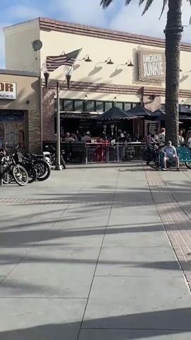 The video opens on a street scene with a building facade in the background. The building has a sign that reads "AMERICAN JUNKIE". There are people seated at outdoor tables in front of the building under umbrellas. Palm trees line the street, and there is a flag flying on a pole. The foreground of the video is a paved walkway with several bicycles parked. The camera pans to the right, revealing more of the street with palm trees and people walking along the sidewalk. The video captures a bustling outdoor scene with a clear blue sky and scattered clouds. There is a sense of a lively atmosphere, possibly a tourist area or a popular gathering spot. People are dressed casually, and the overall impression is one of a sunny day with outdoor activity.