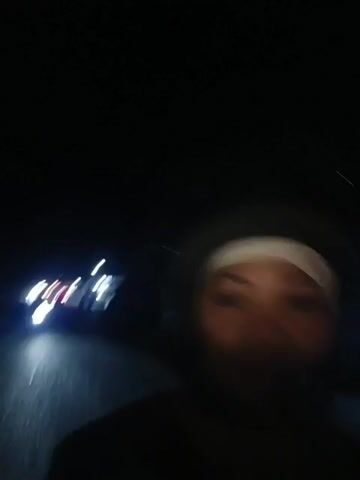 The video begins with a blurry shot of lights on a dark road, possibly from oncoming traffic. The camera then pans slightly to the right, revealing a person's face, partially obscured by a white head covering and a dark helmet. The person appears to be looking forward, and the background continues to show the dark road with streaking lights. The camera movement is unsteady, suggesting it might be mounted on a moving vehicle. As the video progresses, the helmet becomes more prominent, and the person's face is still visible, though the image remains out of focus due to motion blur. The environment is clearly nighttime, with artificial lights from vehicles and possibly streetlights being the primary sources of illumination. The road surface is visible and appears wet or reflective. The video continues with the same blurred perspective, focusing on the person on what appears to be a motorcycle or scooter, traveling at night.