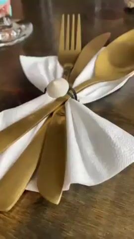 The video shows a close-up of a table setting at a restaurant.  The table is made of dark wood, and there are napkins folded into decorative shapes, some held together by small objects.  There's also gold-colored silverware wrapped in a napkin and held together with a small, white, round item. One napkin holder looks like a piece of driftwood. A person's torso and arm can be seen in the background to the left, and a can of soda is visible behind another cutlery setting. The scene appears to be inside a restaurant, possibly near a window.