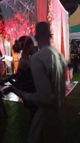 The video appears to be shot at an outdoor event, possibly a party or wedding, with decorated structures in the background. People are moving around and dancing.  A band is playing, with drums visible. Some individuals are dressed in traditional attire. The camera moves erratically, making it difficult to clearly observe the scene.