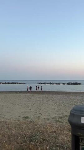 The video shows a group of five people standing on a sandy beach near the water's edge. The sky is a gradient of light blue to pink, suggesting it might be sunset or sunrise. The sea is calm, and a line of rocks is visible further out in the water. The beach has some sparse vegetation growing in the sand, particularly in the foreground. There is a metal object with a circular base in the lower left corner of the frame. The people on the beach appear to be talking or interacting, although specific actions are not easily discernible.