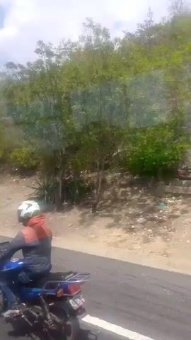 A motorcycle rider is visible in the foreground, wearing a helmet and riding on a paved road. The rider is wearing a gray shirt with red accents. The background features a dirt embankment with vegetation and trees, and a hillside covered in greenery. The video appears to be captured from a moving vehicle, as indicated by the motion blur and perspective.