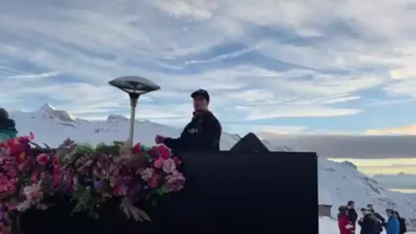 A man in a black jacket and baseball cap is behind a DJ setup decorated with flowers. The DJ setup is on a snowy mountain with a view of other snow-capped mountains in the background. The sky is partly cloudy with long, wispy clouds. There are a few people standing further back, looking at the DJ. A tall, modern lamp stands beside the DJ setup. The DJ is moving his hands as if he is mixing music.