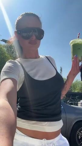 A woman stands outdoors on a bright, sunny day, holding a green drink and car keys, while speaking to the camera. She is positioned in front of a parked grey Mercedes-Benz SUV, with a clear blue sky and green trees in the background. She is wearing a white short-sleeved t-shirt layered under a dark grey or black ribbed camisole, paired with white, low-slung, wide-leg trousers that reveal her midriff. Dark, oversized sunglasses are on her face, and her hair is pulled back. Visible tattoos adorn her arms, and she has light blue nail polish. In her right hand, she holds a clear plastic cup containing a green beverage with a red and white striped straw, along with a set of car keys. She gestures with her left hand as she speaks. A utility pole with a partially visible sign, possibly reading "CYCLE," is seen in the background. The scene suggests a casual, everyday outdoor setting.