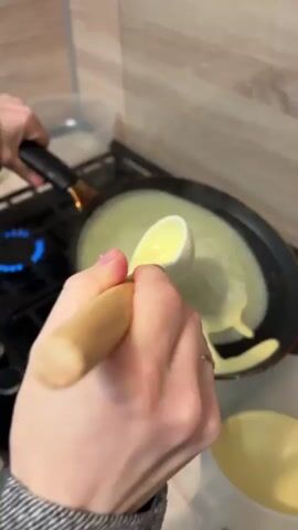 Here is a description of the video:

The video shows a person cooking pancakes on a gas stovetop. The person uses a wooden ladle to pour pancake batter into a non-stick frying pan. They then hold the frying pan by its handle and swirl it to evenly distribute the batter. A blue gas flame burns beneath the frying pan. A blue plate sits to the side with a spatula and two cooked pancakes. A clear container is also nearby, holding more pancake batter.