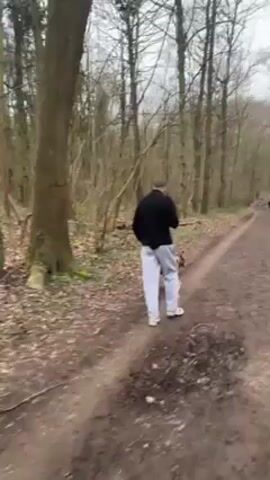 A man walks with his brown dog on a dirt path in a forest. The man is wearing a black shirt and gray sweatpants. The dog is walking near his side. Tall, leafless trees line the path. In the distance, another person is seen walking further down the path. The ground is muddy, and the weather appears overcast.