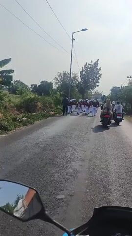 A group of young people in matching uniforms are marching down a road. They are wearing maroon long-sleeved shirts and white pants. Some of them are also wearing white hats. They are walking in a line and appear to be part of a parade or procession. There is a person on a scooter in the background, and the video is filmed from the perspective of someone riding a motorcycle. The road is paved and appears to be in a rural or semi-rural area, with vegetation on the side of the road. The sky is clear and the sun is shining.