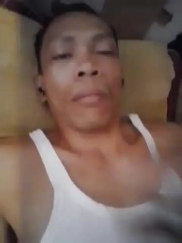 This is a close-up, static video of a person lying down and covering their nose and mouth with their right hand. The person is wearing a white tank top. The background is blurry and appears to be a room with some bedding. The lighting is dim.