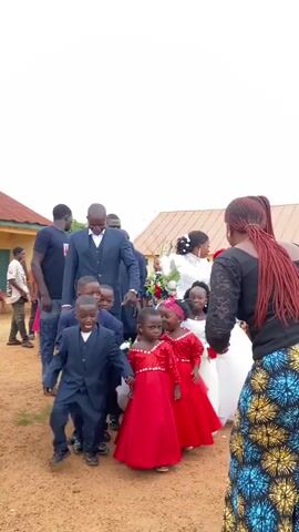 In a medium shot, a wedding party with several children walks across a dirt yard on a cloudy day.  At the front of the group, a man in a navy blue suit smiles, appearing to guide the children.  A line of small boys in matching navy blue suits walks beside him, followed by a line of girls in red dresses.  Behind the girls is the bride, in a white wedding dress.  To the right of the group, a woman in a black lace top and multi-colored skirt reaches out to guide one of the girls.  Behind the wedding party are several onlookers, including a man in a black t-shirt, jeans and a gold chain, and a small child in a green outfit.  The background features a simple building with a rust-colored roof.  The overall atmosphere is festive and joyous.