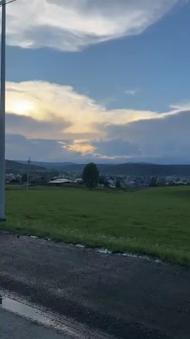 This is a vertical, shaky video of a rural landscape at dusk. The foreground is a dark, gravelly road. Beyond the road, a wide, green field stretches towards a village. The village consists of scattered houses with visible rooftops, nestled amongst trees and rolling hills in the distance. A lone tall tree stands in the middle ground. The sky above is a mix of blue and dramatic, cloud-streaked sunset colors, with the sun partially obscured by clouds, casting a golden glow. A thin wire is visible diagonally across the upper portion of the frame. There are no people or discernible actions captured in the video. The overall impression is one of a quiet, pastoral evening.