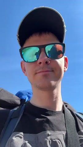 The video shows a man walking on a dirt road. The man is wearing a black t-shirt, a backwards grey and white baseball cap, and blue-tinted sunglasses. He is carrying a large blue backpack. The background consists of greenery, fences, and some residential buildings. The sky is blue. The camera appears to be hand-held. The man seems to be taking a selfie video. He smiles at the camera.