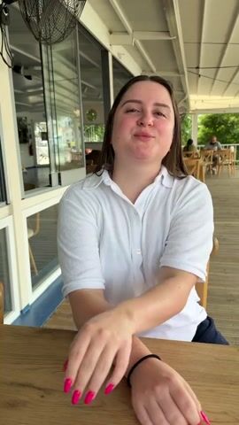 The video opens with a medium shot of a young woman sitting at a wooden table in an outdoor restaurant. She is wearing a white collared shirt and has her arms crossed. She smiles at the camera and then turns her head to look off to the side. The camera then pans to the right, revealing an empty outdoor dining area with more tables and chairs. Beyond the dining area, there is a scenic view of a body of water with several boats docked and a lush green hillside in the background. The video ends with a final shot of the scenic view.